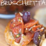 a crossonti with pepperoni, kalamata olives, parmesan cheese on wood cutting board with glass bowl of bruschetta