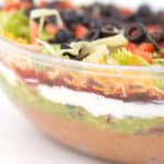 a close up of a clear bowl with layers of beans, guacamole, salsa, sour cream, lettuce, cheese, tomatoes, and olives