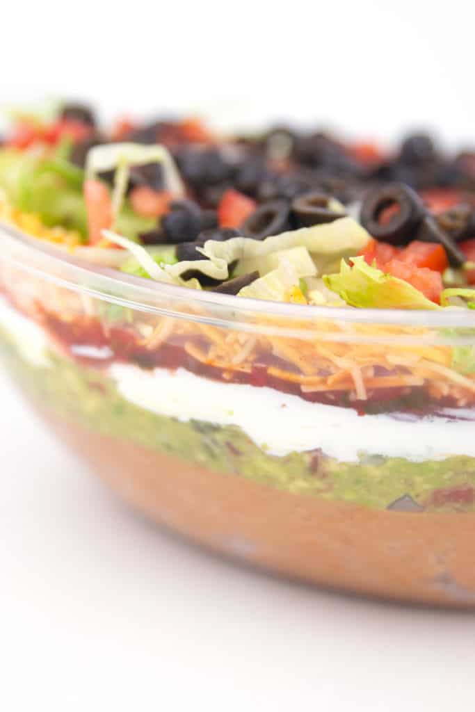 a close up of a clear bowl with layers of beans, guacamole, salsa, sour cream, lettuce, cheese, tomatoes, and olives