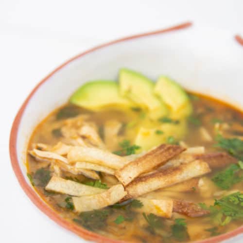 a white bowl of mexican chicken tortilla soup with avocados, tortilla stripes and cilantro