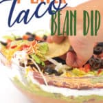 A clear bowl with layers of beans, guacamole, salsa, sour cream, lettuce, cheese, tomatoes, and olives with a chip taking a dip