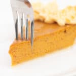 a piece of pumpkin pie on a white plate with a fork piercing it