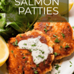 a white plate of crispy salmon patties with a white sauce on top and green lettuce and lemon