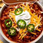 white bowl of turkey chili with shredded cheese, sour cream and jalapenos on top with crackers on the side