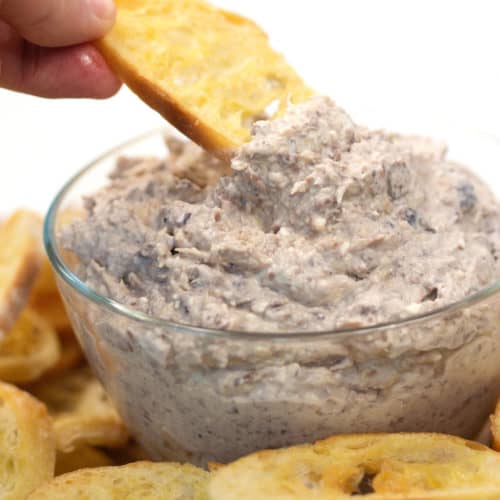 a glass bowl of whipped cheese with olives and artichokes and toasted bread dipping