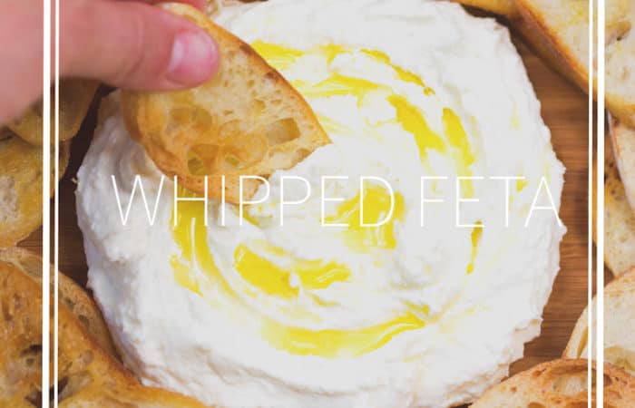 crostinis on a wood cutting board dipping into whipped feta with olive oil drizzle
