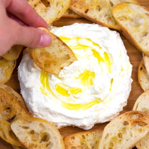 crostinis on a wood cutting board dipping into whipped feta with olive oil drizzle