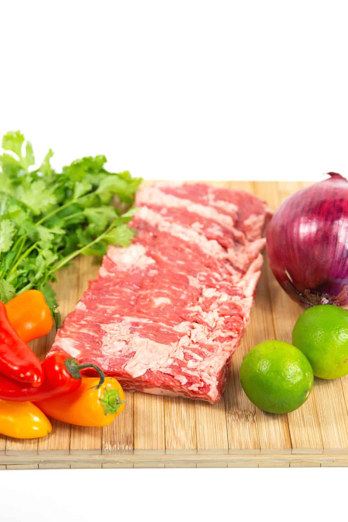 wood cutting board of raw flank steak, whole onion, peppers, cilantro, and lime