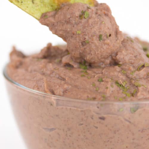 a glass bowl of black bean dip with parsley flakes on top with a green tortilla chip dipping