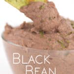 a glass bowl of black bean dip with parsley flakes on top with a green tortilla chip dipping