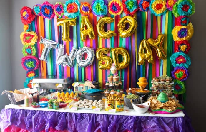 streamer back drop with mexican flowers border wih taco twosday balloons with mexican food table