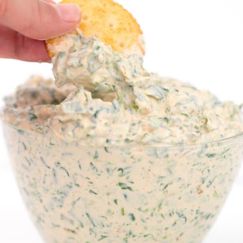 a glass bowl with spicy ranch spinach dip in it dipping a cracker in the dip
