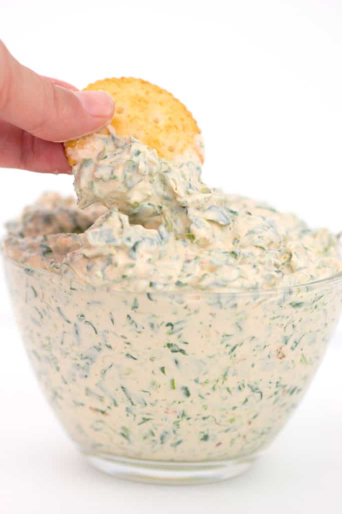 a glass bowl with spicy ranch spinach dip in it dipping a cracker in the dip