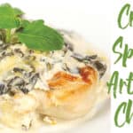 white plate of a browned chicken breast with creamy spinach and artichoke sauce with basil leaves on top