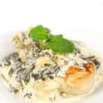 white plate of a browned chicken breast with creamy spinach and artichoke sauce with basil leaves on top