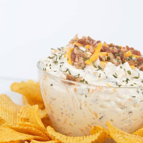glass bowl of easy cheddar bacon ranch dip with cheese and bacon on top with chips around the bowl