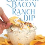 dipping a chip into a glass bowl of easy cheddar bacon ranch dip with cheese and bacon on top with chips around the bowl