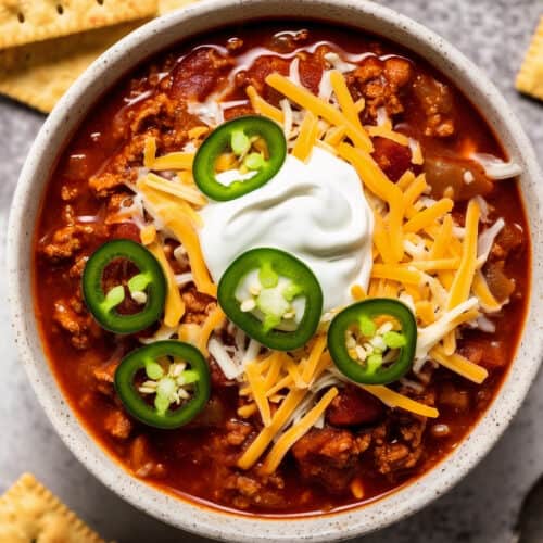 white bowl of turkey chili with shredded cheese, sour cream and jalapenos on top with crackers on the side