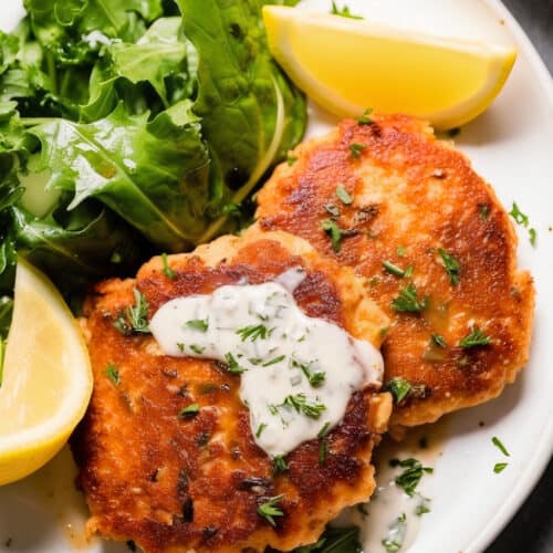 a white plate of crispy salmon patties with a white sauce on top and green lettuce and lemon